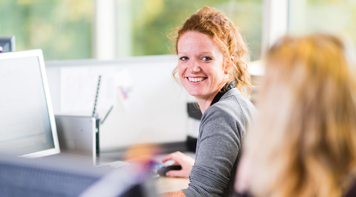 Woman smiles while conversing with a colleague at her desk.